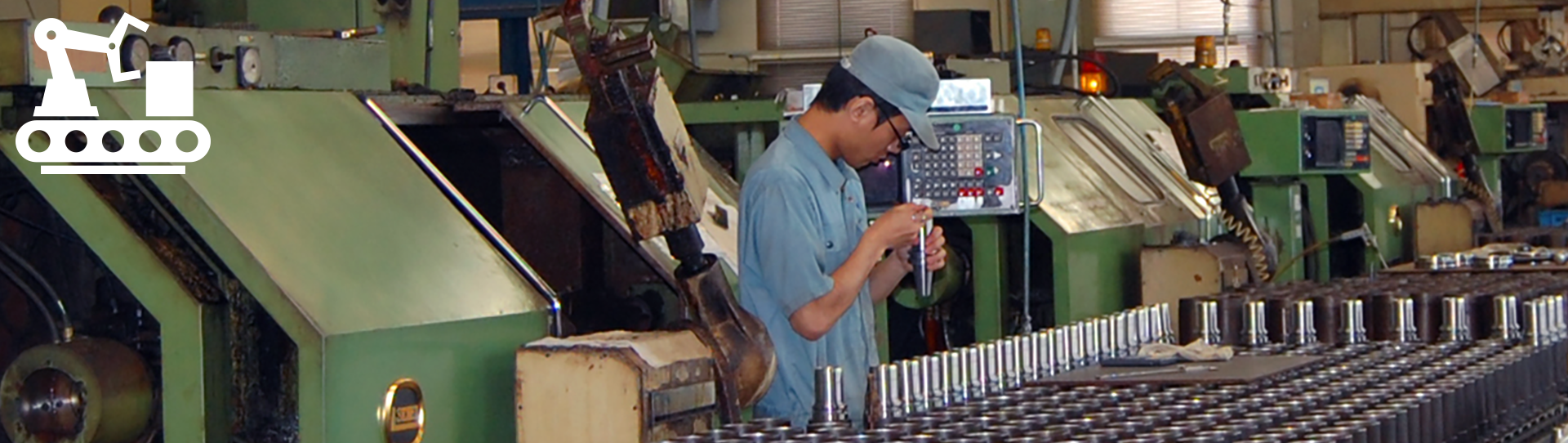 A worker wearing a cap and uniform inspects a metal part in a factory with green industrial machines and rows of cylindrical components; a white machinery icon appears in the top left corner.