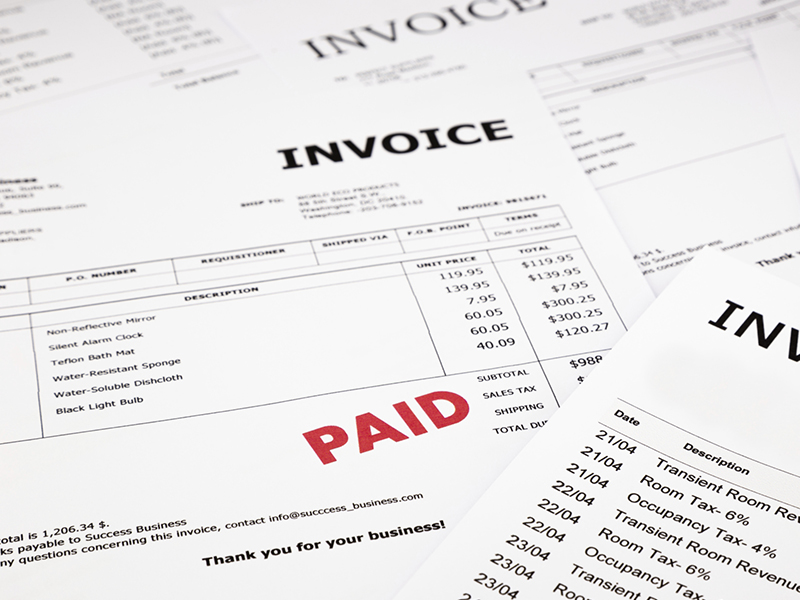 Accounting_800x600 Several invoices are spread out on a surface. One invoice shows a large red “PAID” stamp across it, while others display itemized lists, prices, and company contact information.