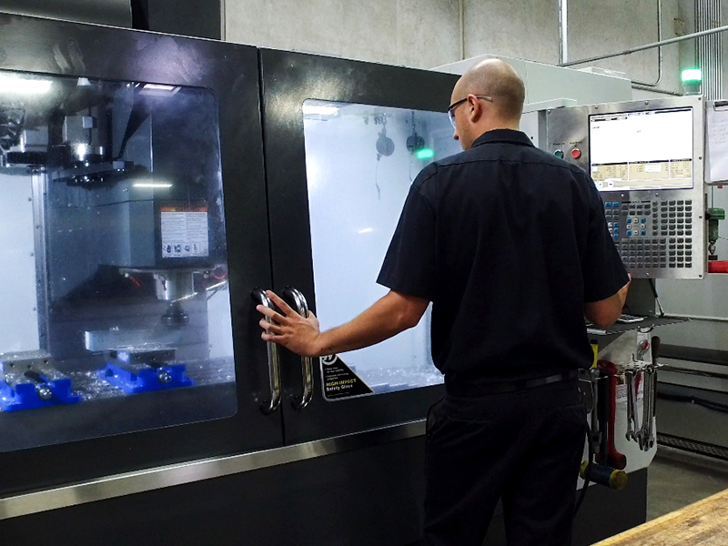 Engineering_800x600 A person in dark clothing operates a large CNC machine in an industrial setting, with their hand on the machine door and control panels visible in the background.