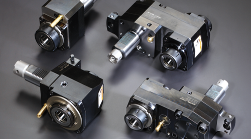 Four industrial metal tool holders with cylindrical and rectangular components are arranged on a dark surface. Each piece features various fittings, bolts, and metallic finishes, with some having attached brass-colored connectors.