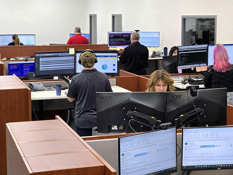 Inside-Sales-Office_800x600 Several people work at computer desks in an open office. Some are seated while others stand, facing large monitors. The workspace has wood-paneled dividers, and most desks have multiple screens and office equipment.