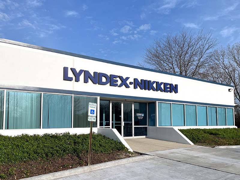 A single-story commercial building with large windows and a sign reading LYNDEX-NIKKEN above the entrance. There are bushes, a concrete walkway, and a handicapped parking space in front. The sky is clear.