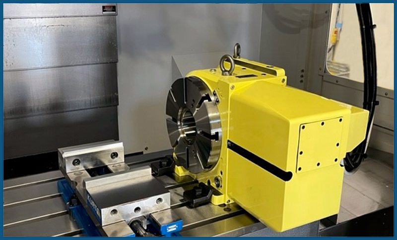 RT-Integration-left_800x485 A yellow rotary table is mounted on the bed of a CNC machine next to a metal vise. The table has a shiny circular faceplate and is positioned for machining operations.