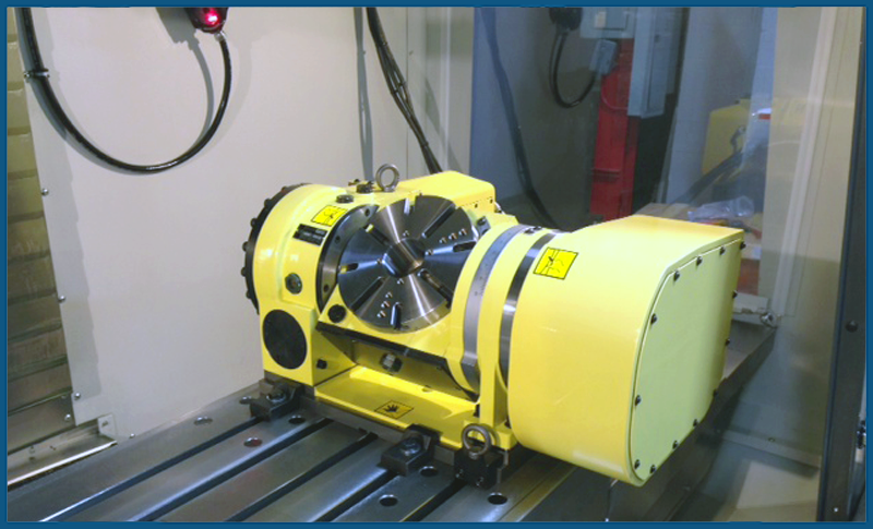 RT-Integration-right_800x485 A yellow rotary table attachment is mounted on the bed of a CNC milling machine inside an enclosure, with metal rails underneath and control wiring visible in the background.