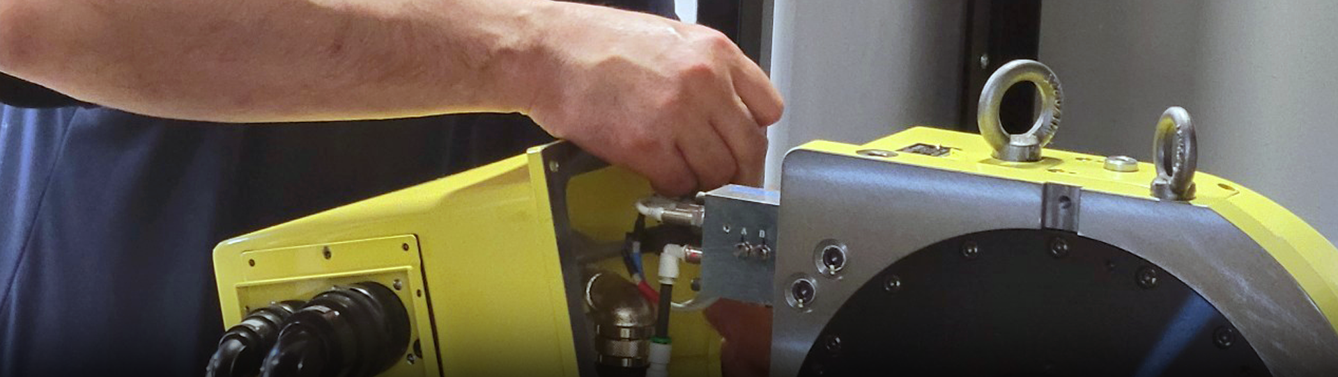 A person’s hand adjusts a component on a large yellow and silver industrial machine with cables and metal fittings visible.