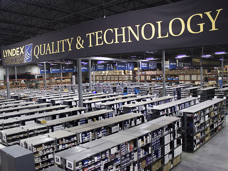 Warehouse-Overhead_800x600 A large warehouse with rows of shelves filled with boxes and bins, overhead lighting, and a prominent sign reading LYNDEX NIKKEN QUALITY & TECHNOLOGY hanging from the ceiling.