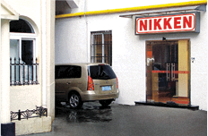 A tan minivan is parked next to a building with a glass door and a sign above it that reads NIKKEN in red letters. A white building with a window and black railing is visible on the left.
