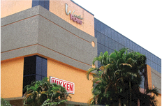 A modern commercial building with an orange and gray facade, large windows, and palm trees in front. The building has a visible “NIKKEN” sign and another sign reading World Trade Center.