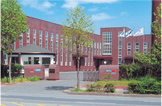A red-brick building with large windows and several flags on poles out front, surrounded by trees and greenery, seen from across a street on a clear day.