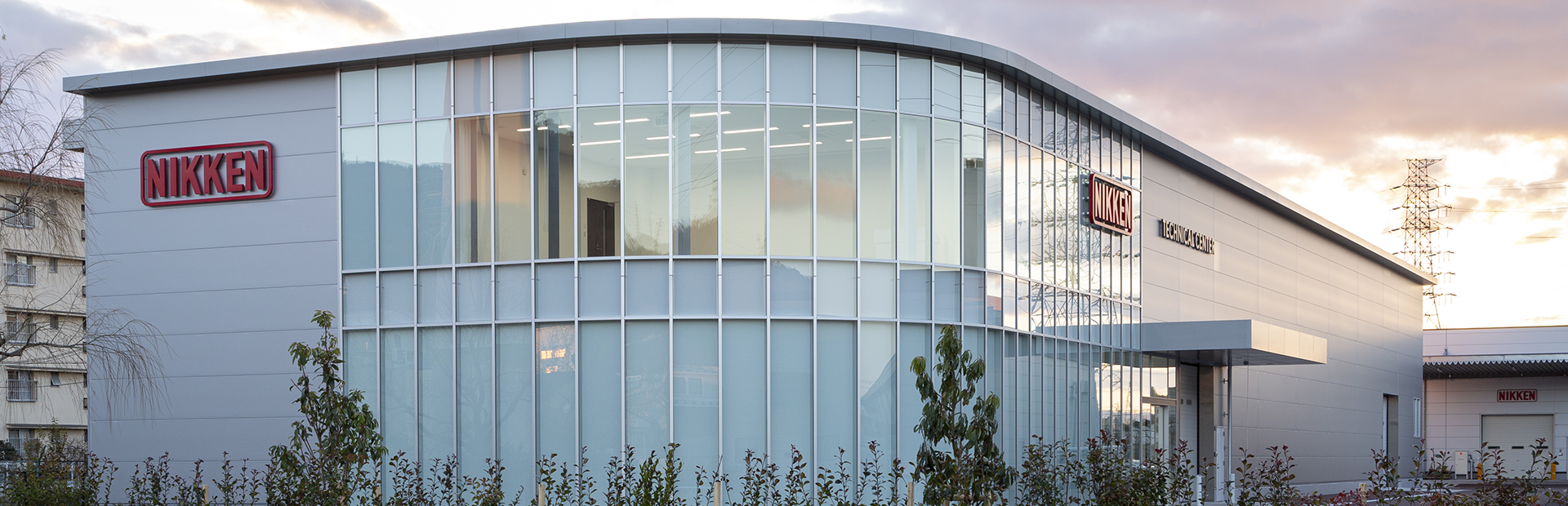 About-Us-Nikken-Building_1920x620 A modern, two-story building with large glass windows and a curved facade features red Nikken logos. The setting is outdoors with some greenery and power lines visible in the background.