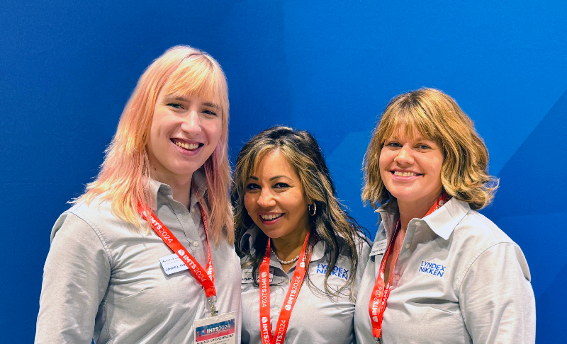 Three people wearing gray shirts with LYNDEX NIKKEN logos and red lanyards stand together and smile in front of a blue background.