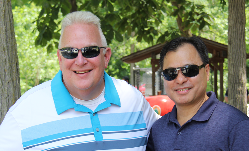 Two men wearing sunglasses and collared shirts stand side by side outdoors. Trees, green leaves, and a wooden shelter are visible in the background. Both men are smiling at the camera.