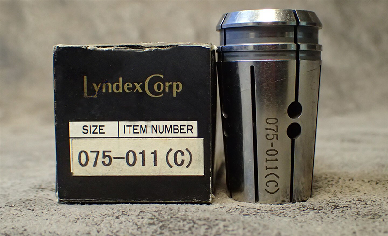 A metal collet marked 075-011(C) is positioned next to a black and white Lyndex Corp box labeled with the same item number and size information. Both items are on a textured gray surface.