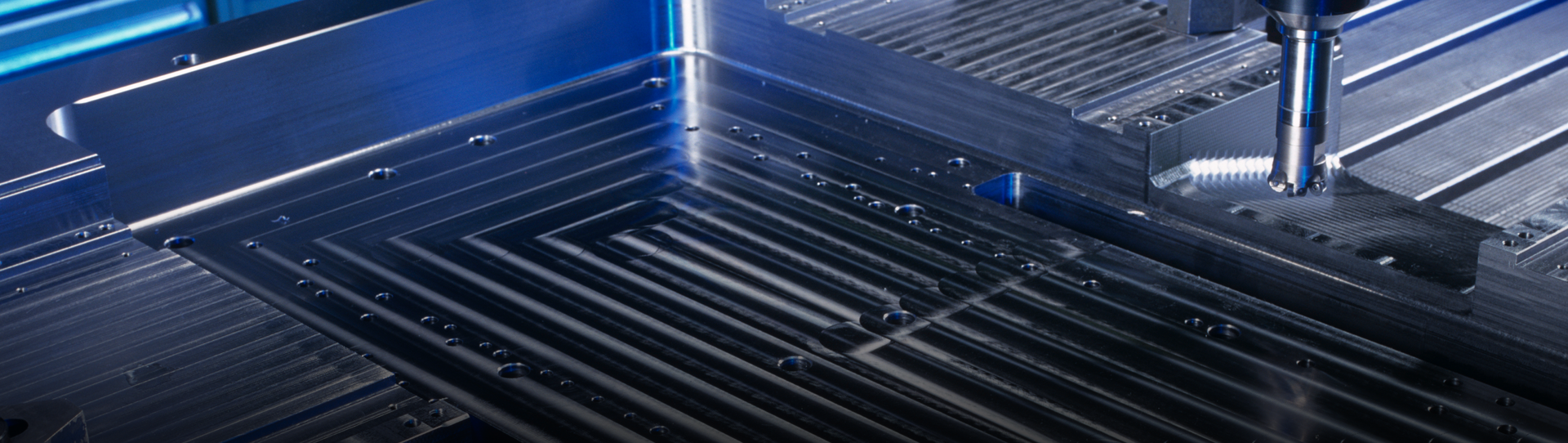 A close-up view of a CNC milling machine cutting precise grooves into a flat metal surface, with visible tool marks, metal chips, and a blue-tinted lighting.