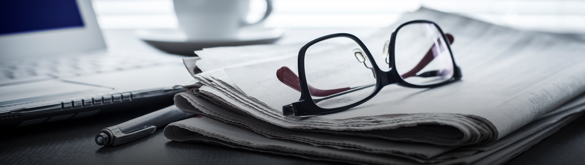 News-Media-Hero_1920x542 A pair of black-rimmed eyeglasses rests on a folded newspaper next to a pen, with a laptop and a cup in the background.