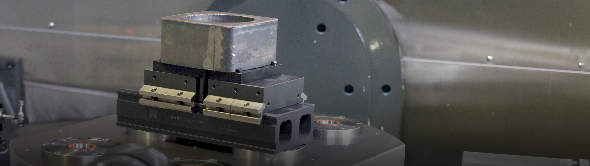 A rectangular metal block is secured in a precision vise on a machine tool table, set against an industrial background with metal surfaces and mechanical components visible.