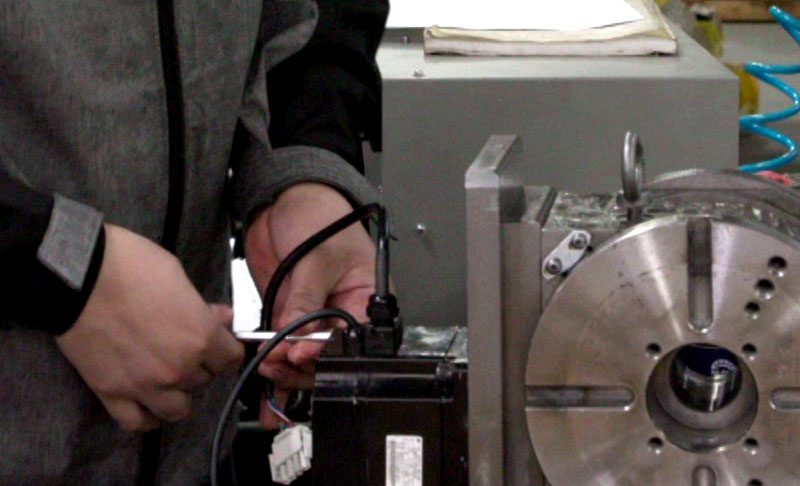 A person in a gray jacket is adjusting cables on a black box mounted on a Rotary Table.