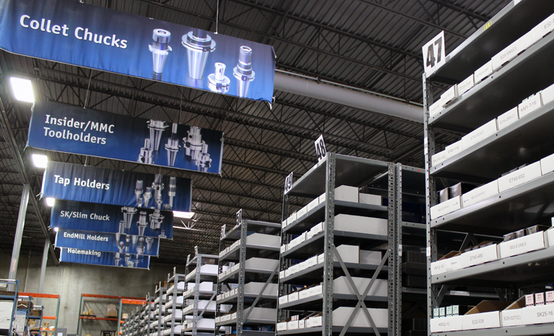 Warehouse interior with tall metal shelves holding white boxes, labeled signs hanging from the ceiling for collet chucks, toolholders, and tap holders.