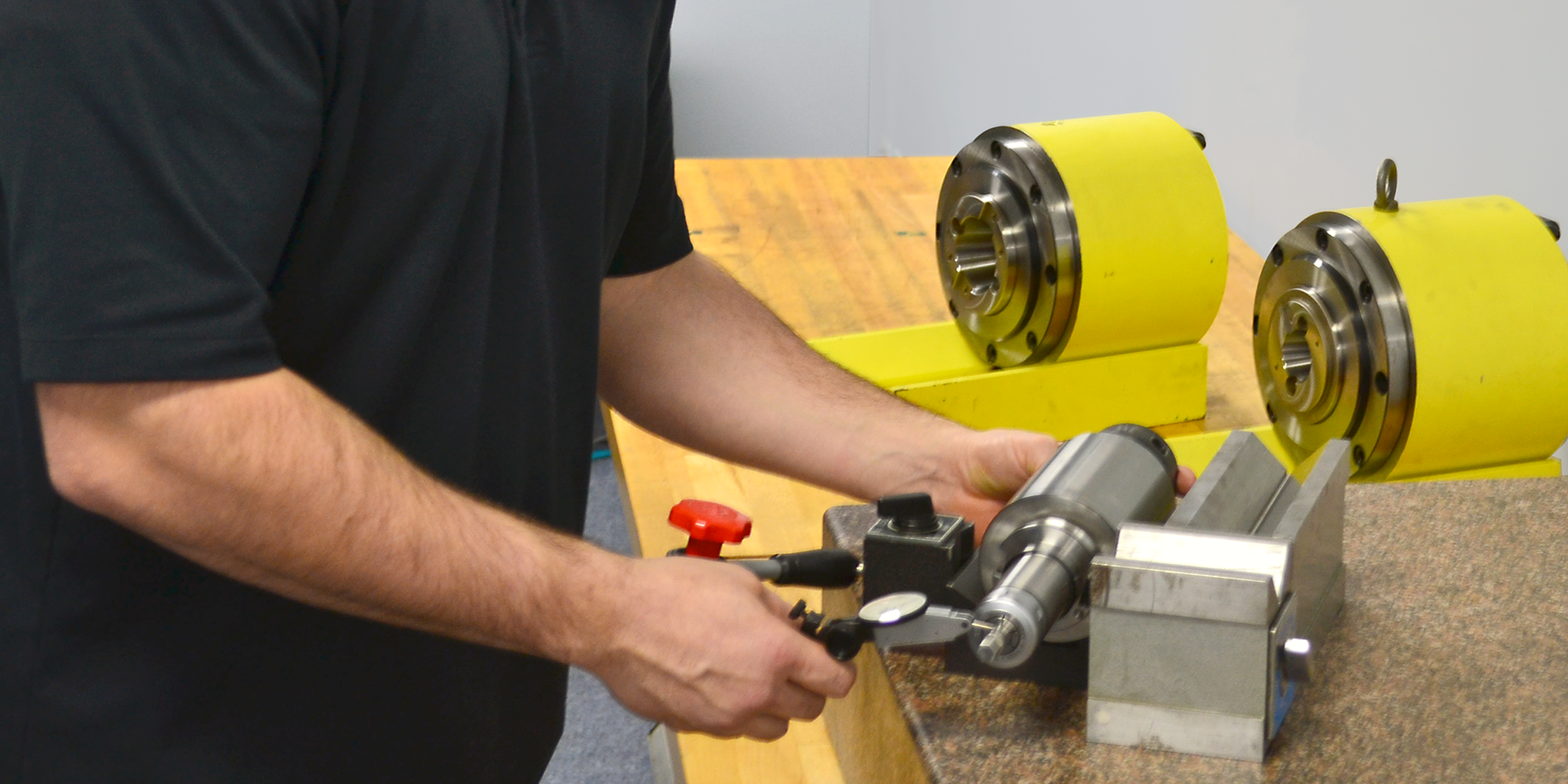 Factory-Trained-Service-Blog-Banner-2000x1000 A person in a black shirt uses a dial indicator to measure a cylindrical metal part held in a vise, with two yellow industrial components in the background on a wooden surface.