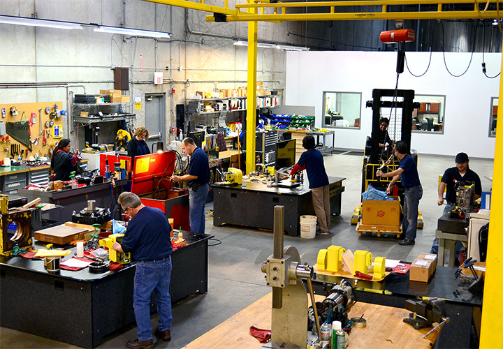 ServiceDSC_0052-2 Workers in a busy industrial workshop use tools, assemble machinery, and operate equipment at various stations. The space has concrete walls, shelves with parts, and overhead yellow safety rails.