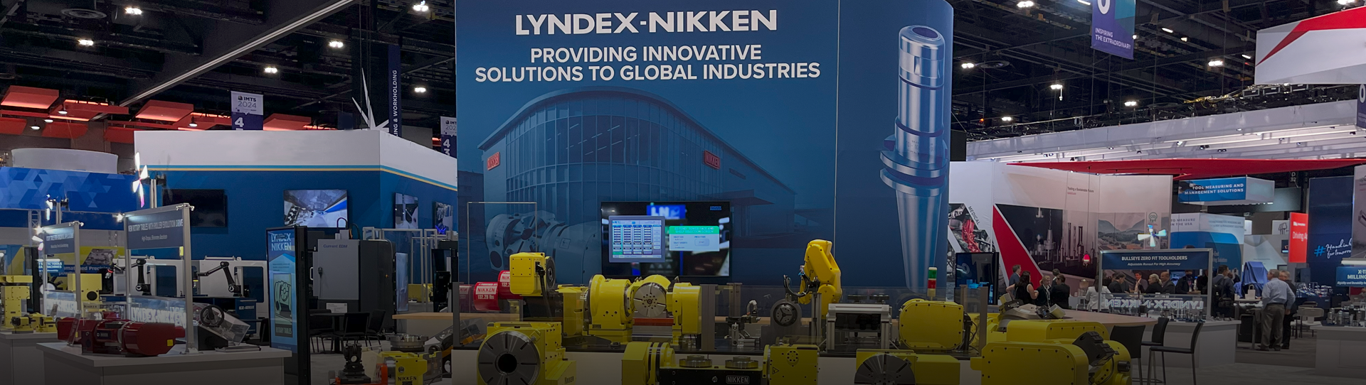 A trade show booth for LYNDEX-NIKKEN displays industrial equipment, including yellow robotic arms and machinery, with a large blue backdrop featuring the company name and slogan about providing solutions to global industries.