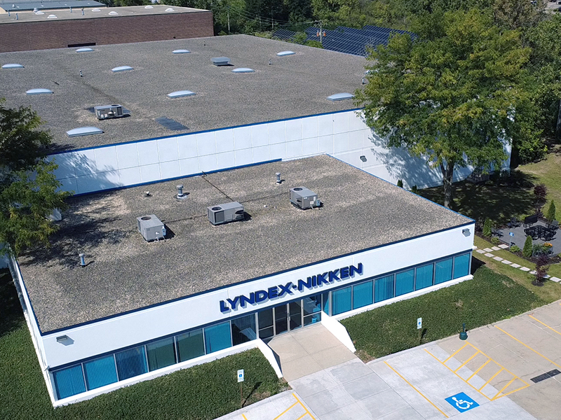 Aerial view of a single-story commercial building with LYNDEX-NIKKEN signage above the main entrance. The building has a flat roof, blue-trimmed windows, and nearby parking spaces, including a marked accessible spot.