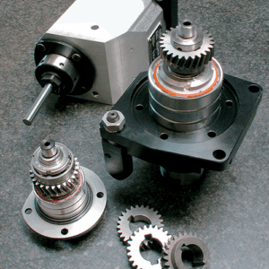 Three metal gear assemblies and several loose gears are displayed on a gray surface. One assembly is attached to a right angle head, and the others are disassembled with visible gear components..