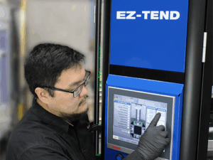 A man wearing glasses and gloves operates a touchscreen control panel on a blue machine labeled EZ-TEND in an industrial setting.