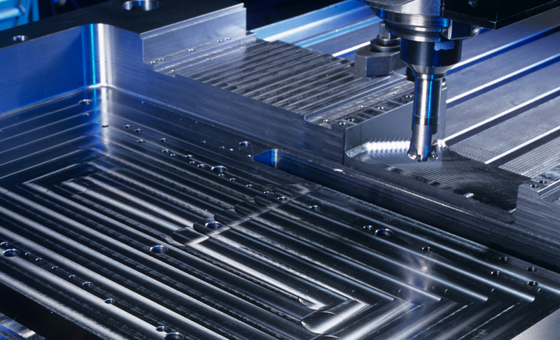 Resources-Search-Application_800x486 A CNC milling machine drills into a flat, rectangular block of metal, creating precise grooves and patterns on its surface. The metal appears shiny and is clamped securely.