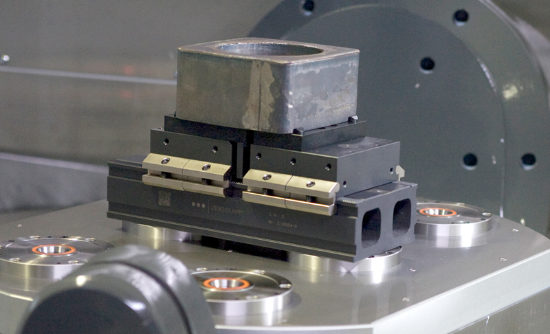Resources-Search-Product_800x486 A metal workpiece is secured in a precision work holding vise, which is mounted on a CNC machining table with circular positioning fixtures. The setup is prepared for a machining or milling process.