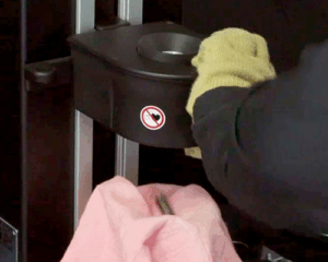 A person wearing thick yellow gloves is holding a metal object near a shrink fit machine inductor. A pink cloth covers part of the scene.