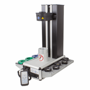 A Start 2 Shrink Fit Machine with a vertical column, adjustable arm, cylindrical holder marked with a no-hand symbol, several multicolored rings, and a connected remote control rests on a white background.