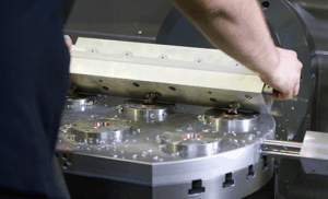 A person positions a large metal component onto a machine with circular fixtures and metal shavings visible, utilizing a clamping system in a manufacturing or machining environment.
