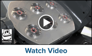 A metal workholding fixture with six circular mounts is shown, highlighting its clamping system. A play button overlay appears in the center. The word “Workholding” and an icon are on the left, with “Watch Video” text at the bottom.