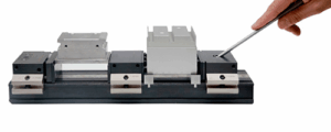 A hand uses a tool to adjust a metal fixture featuring a precise clamping system that holds three different metal components, all mounted on a black rectangular base against a white background.