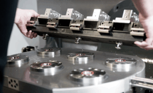 Two people are installing or removing a precision mechanical fixture with several adjustable clamps onto a round metal platform containing multiple circular holders in a manufacturing or laboratory setting.