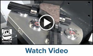 A metal workpiece is clamped onto a round CNC machine fixture with a robust clamping system, featuring multiple chucks and rails. There is a workholding icon in the lower left corner and a play button in the center of the image.