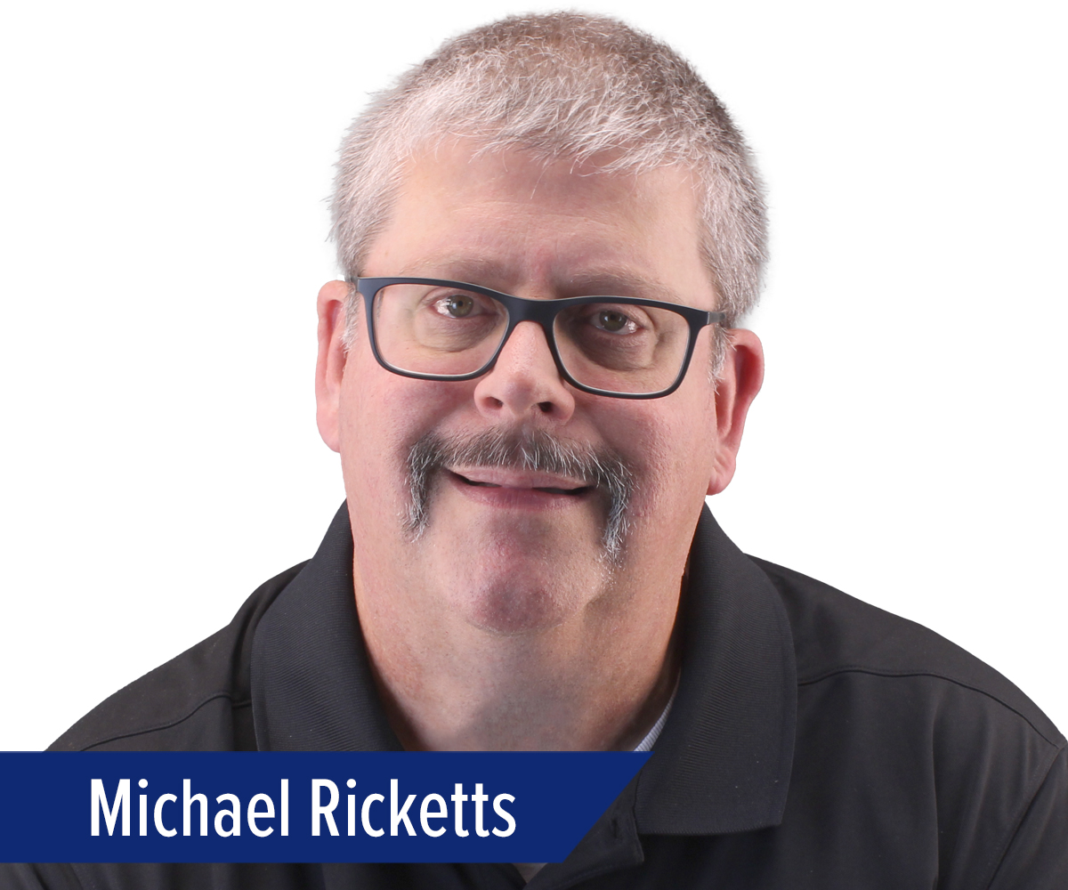 Michael Ricketts_Press Release Profile_300dpi A middle-aged man with short gray hair, glasses, and a mustache is wearing a black collared shirt. The name Michael Ricketts appears in white text on a blue banner at the bottom left.