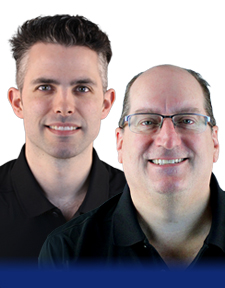Two men, both wearing black collared shirts, stand side by side and smile at the camera. One has short dark hair, the other has glasses and is balding. The background is plain white with a blue gradient at the bottom.