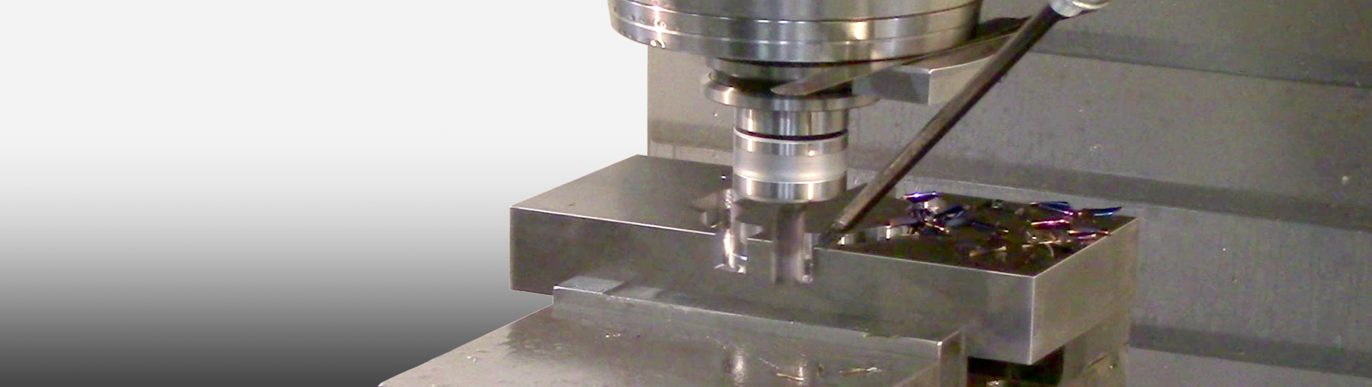 A CNC milling machine is cutting a rectangular metal block, with metal shavings visible on the blocks surface. The surroundings are metallic and industrial in appearance.
